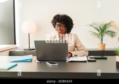 Focused and determined African American woman dressed in a suit, engaged in important work on her laptop at her desk while surrounded by office Stock Photo