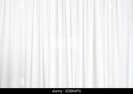 A long white curtain hangs in front of a white background. The curtain is made of a sheer material, allowing light to pass through it. The curtain is Stock Photo