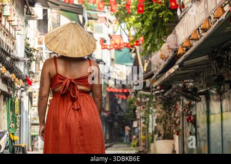 View of a woman in an orange dress and conical hat walks down a narrow alleyway adorned with red lanterns, casting a warm glow, Hanoi, Hanoi, Vietnam. Stock Photo
