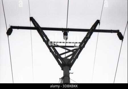Black and white film photograph of a high-voltage electricity pylon silhouette against a pale overcast sky, viewed from below. Minimalist composition emphasizing geometric structure and power infrastructure Stock Photo