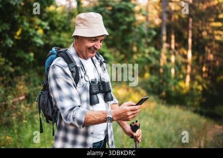 Portrait of smiling senior hiker with backpack and binoculars using smartphone while discovering nature during solo mountain hike. Stock Photo