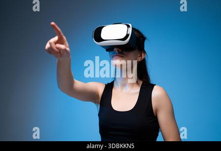 Woman in VR headset pointing forward while exploring immersive virtual reality world in blue studio Stock Photo