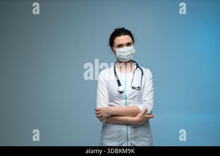 Confident female doctor in medical mask with stethoscope posing with folded arms on blue background Stock Photo