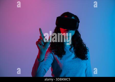 VR headset woman interacts with invisible virtual interface reaching forward in mixed neon red blue lighting Stock Photo