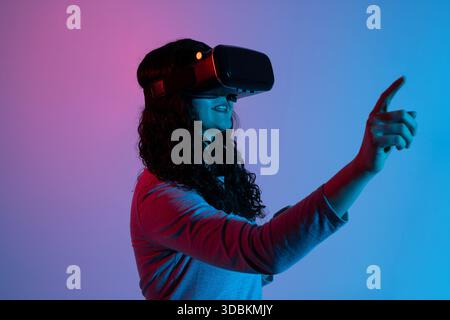A woman wearing a virtual reality headset reaches forward and interacts with digital elements in soft neon blue and pink lighting. Stock Photo