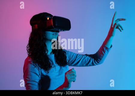 A woman immersed in virtual reality raises her hand toward digital space in neon light Stock Photo