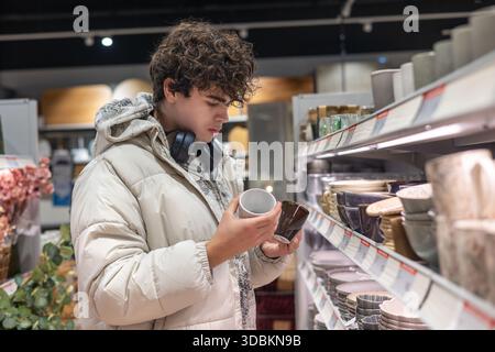 Teenager Comparing Mugs in Kitchenware Aisle – Young Customer Choosing Ceramic Cups in Home Goods Store. Stock Photo