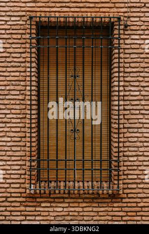 Window with a protective wrought iron grate and closed wooden blinds on an old brick wall, reflecting typical architecture in Cordoba, Andalucia, Spai Stock Photo
