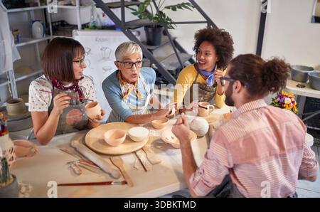 Elderly teacher explaining to student on handmade pottery worhshop while others painting pot with brush.Creative, hobby ,activities concept Stock Photo