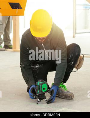 Construction worker wearing protective workwear and yellow hardhat using rotating laser level on tripod, taking measurements at construction site with bright sunlight Stock Photo
