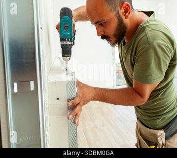 Concentrated construction worker carefully installing metal corner bead on drywall with cordless screwdriver during renovation or construction work in an apartment Stock Photo