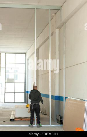 Construction worker installing aluminum pillars for the construction of a plasterboard wall in an industrial warehouse, using protective gloves and with his helmet hanging from his trousers Stock Photo