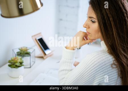 beautiful young woman in a white sweater sits at a work table with a pensive view close-up || Model released Stock Photo