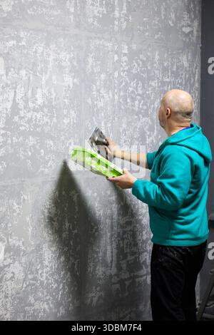 In a medium shot, a plasterer is captured applying decorative plaster to a wall with a trowel. The man, bald with a visible beard, is dressed in a tur Stock Photo