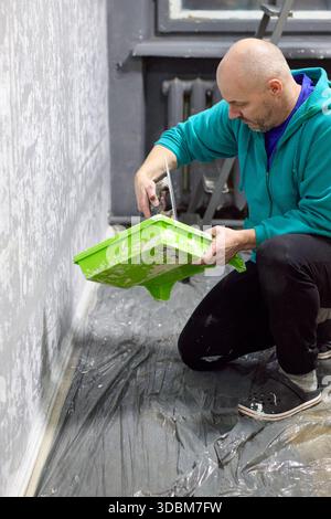 This image features a dedicated craftsman in the process of applying textured plaster to a gray wall indoors. Kneeling on a plastic-covered floor, he Stock Photo