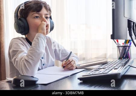 A child aged 7-9 sits at home behind a computer, writing assignments in a notebook. The image captures remote learning, digital study routines, challe Stock Photo