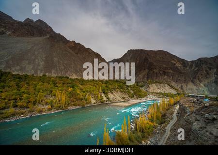 View of the vibrant turquoise river winding through the rugged mountains, with golden trees lining the banks, contrasting against the grey sky, Gupis, Stock Photo