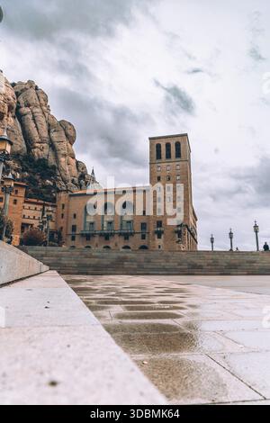 View of the monastery standing tall against the backdrop of rugged mountains under a moody sky, with reflections on the ground, Montserrat, Catalonia, Stock Photo