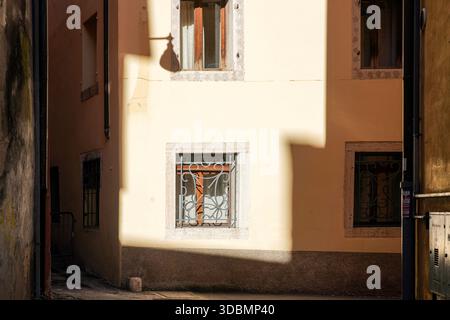 Belluno, Italy, Afternoon sun illuminates a narrow alleyway, casting sharp shadows on pale yellow walls of a building in the Dolomites town, ornate iron grill over the lower window, shadow of an overhead lamp on the stucco surface Stock Photo