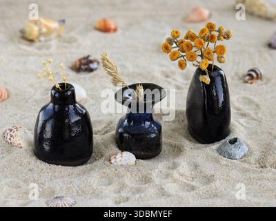 Three small handmade ceramic vases with dried flowers arranged on natural sand with seashells. Minimalist still life composition inspired by coastal a Stock Photo