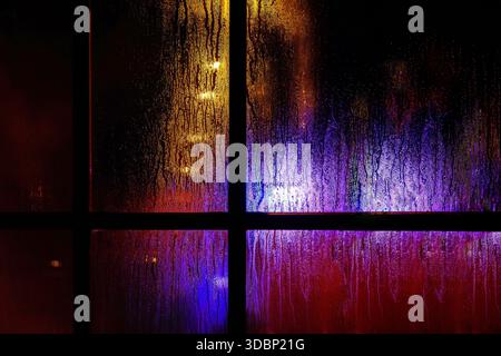 A dark window pane with rain droplets at night. Blurred yellow and blue festive christmas lights through the wet glass covered with condensate drops. Stock Photo