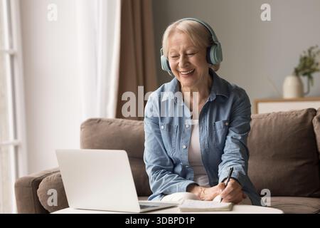 Aged lady in headphones learning online using laptop taking notes Stock Photo
