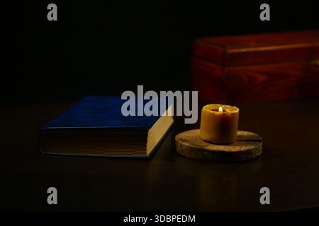 Burning wax candle with spell book and old wooden box on the wood table on dark black background with empty space. Ancient old magic objects Stock Photo