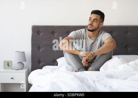 Thoughtful Young Arab Man Sitting on Bed in a Comfortable Room Contemplating Life Stock Photo