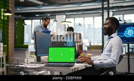 Technician next to office coworkers developing software applications using green screen laptop, integrating AI automation. IT expert debugging applications with mockup notebook, camera A Stock Photo