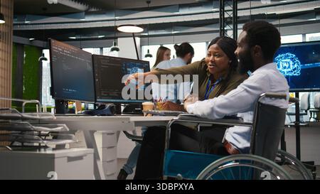 Smiling startup engineer and colleague with disability brainstorming, using machine learning tech. Happy african american man in wheelchair and coworker in office using AI predictive models, camera A Stock Photo