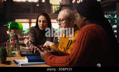 Mature academic colleagues prepare documents for university coursework or exams, transcribing essential ideas from textbooks. Research and education in a quiet public library. Stock Photo