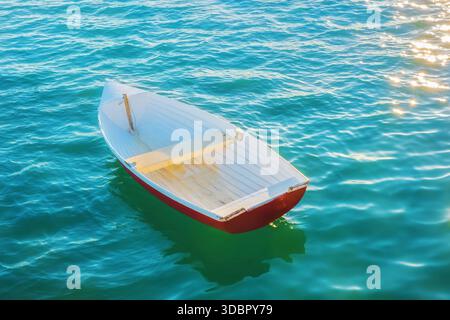 A white and red rowboat drifts peacefully on tranquil water, surrounded by the expanse of bright, blue sea Stock Photo