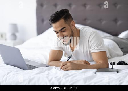 Young Arab Man Studies Online in Bed, Taking Notes on Distance Learning While Smiling Stock Photo