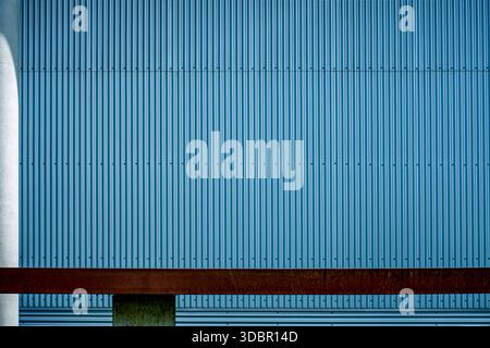 A rusty brown metal bench sits in front of a blue, corrugated metal facade in Augsburg, Germany. The vertical lines of the wall contrast with the horizontal bench and a white concrete pillar on the left. Stock Photo