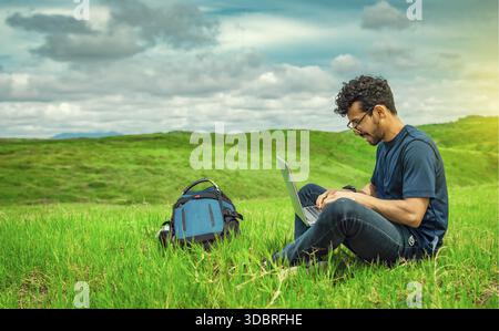 Smiling freelancer young man with laptop working from the field. Close up of man sitting on green grass working with his laptop. Smiling man with lapt Stock Photo