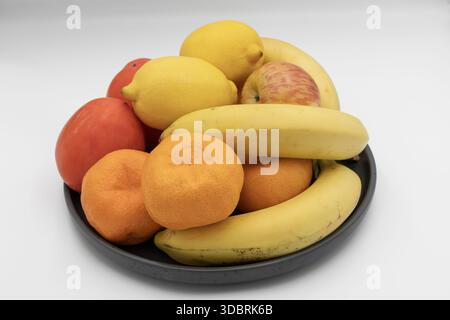Fresh assorted fruits in a black bowl with bananas, lemons, oranges, apples and persimmons isolated on white background Stock Photo