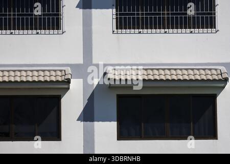 Minimalist exterior of white modern building features dark window glass with metal grille and strong shadow on wall creating abstract geometric patter Stock Photo