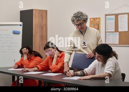 Middle aged Caucasian woman instructing three young adult women of diverse ethnicities wearing prison uniforms sitting at table, completing written assignments during prison education class Stock Photo