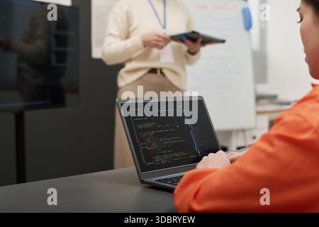 Young adult Caucasian woman sitting at desk using laptop with programming code on screen, while middle aged woman standing in background holding tablet during prison education class Stock Photo