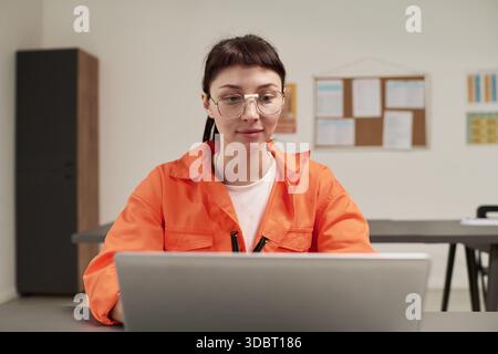 Caucasian young adult woman wearing glasses sitting at desk using laptop computer in classroom setting participating in prison education program with focus on learning Stock Photo