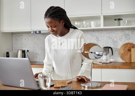 Young Black Woman Using Laptop in Kitchen While Smiling and Leaning Forward Stock Photo