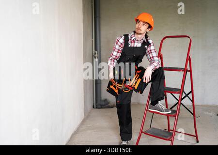 Pensive male laborer, builder or repairman ready for repairing or building, wears overalls and protective helmet, works at construction site stands on Stock Photo