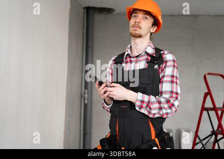 Builder, foreman, repairman wearing tool belt hard hat and overalls holding smartphone looking on his work at construction site. copy space Stock Photo