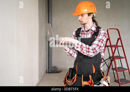 Portrait of young builder, foreman or repairman in orange helmet with laptop indoors Stock Photo