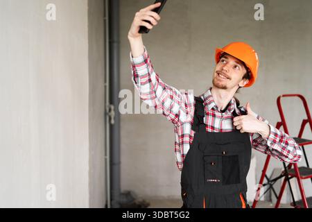 Builder, foreman, repairman wearing tool belt hard hat and overalls at construction site making point of view photo showing thumb up, like gesture. Ba Stock Photo