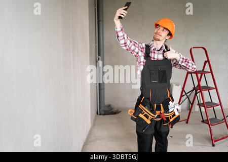 Builder, foreman, repairman wearing tool belt hard hat and overalls at construction site making point of view photo showing thumb up, like gesture. Ba Stock Photo