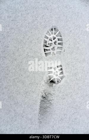 Single deep boot sole tread imprint in fine fresh snow, vertical top-down view, white granular surface, dark rubber pattern visible, overcast daylight Stock Photo