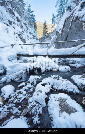 View of icy stream flows under a snow-laden wooden bridge, framed by snow-covered cliffs and evergreen trees against a bright sky, Prosiek, Zilinský k Stock Photo