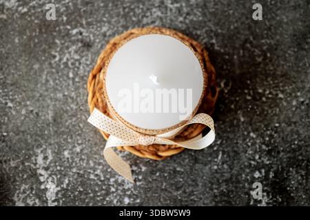White candle wrapped in rustic burlap and decorated with beige ribbon, placed on woven mat over dark textured background. Minimalist cozy decor for ho Stock Photo