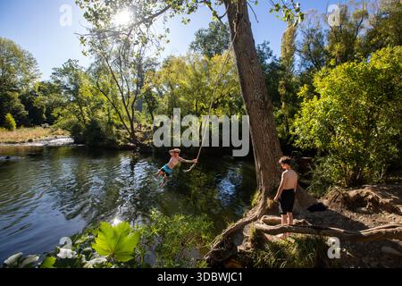 View of a boy swinging on a rope swing over the reflective waters of a river surrounded by lush green trees, Maitai Valley Road, Nelson, Nelson Region Stock Photo